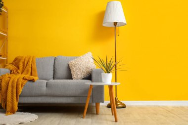 Ekose ve yastık gri koltukta, masa ve lamba sarı duvarın yanında.
