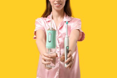 Beautiful young woman with oral irrigator and electric toothbrush on yellow background