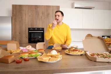 Mutfaktaki sağlıksız yiyeceklerle dolu masada oturan genç adam. Aşırı yeme kavramı