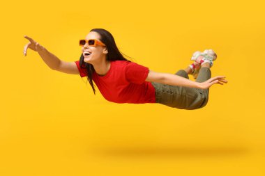 Güneş gözlüklü neşeli genç kadın uçuyor ve sarı arka plandaki bir şeyi işaret ediyor.