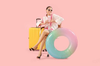 Happy female office worker in sunglasses sitting on armchair near inflatable ring and suitcase on pink background. Summer vacation concept