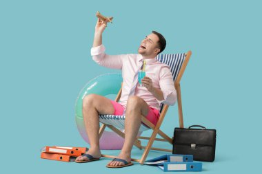 Happy male office worker sitting on deckchair with cocktail and toy plane on blue background. Summer vacation concept