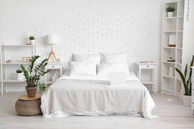 Interior of light bedroom with houseplant and laptop on white bed