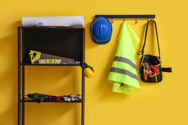 Güvenlik yeleği, inşaat aletli raf ünitesi ve sarı duvarın yanında şapka.