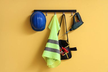 Sarı duvarda asılı çelik yelek, şapka ve aletler.