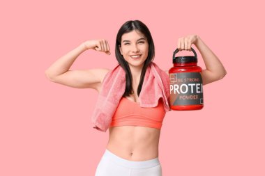Sporty young woman with bottle of protein powder showing muscles on pink background