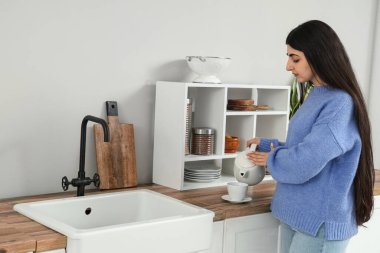 Pretty young woman pouring tea into cup in light kitchen