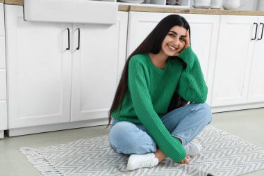 Pretty young woman sitting on rug in light kitchen