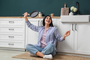 Pretty young woman with frying pan sitting on rug in modern kitchen