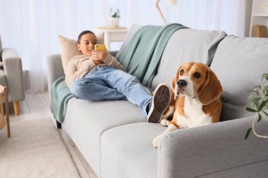 Little African-American boy with mobile phone and cute beagle dog lying on sofa at home