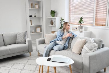 Young woman sitting on sofa and holding cup in light living room