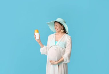 Mavi arka planda güneş kremi şişesi olan güzel hamile bir kadın.