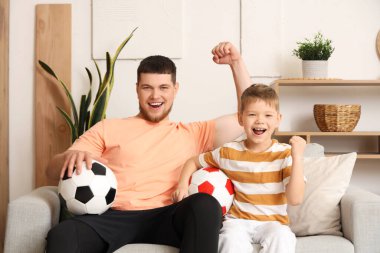 Genç adam ve küçük oğlu evde futbol toplarıyla futbol takımı için tezahürat yapıyorlar.