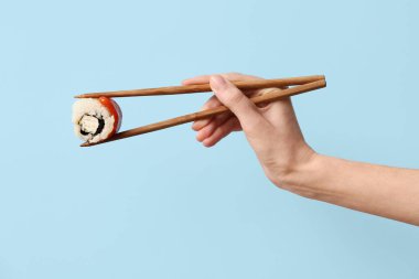 Hand with tasty sushi roll and chopsticks on blue background