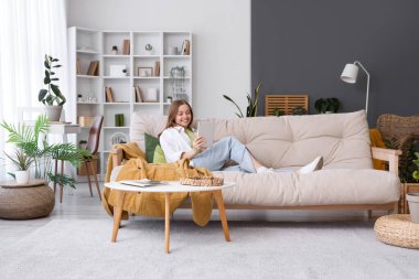 Evdeki kanepede tablet bilgisayarlı genç bir kadın yatıyor.