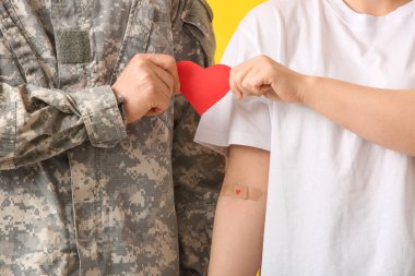 Blood donor with applied medical patch and soldier holding paper heart on yellow background