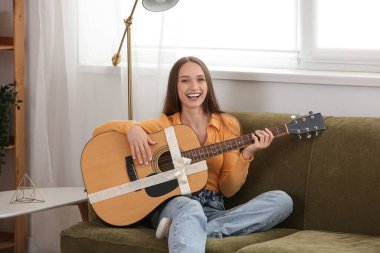 Happy young woman with gift guitar sitting on sofa at home