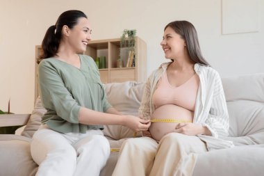 Young pregnant woman with doula measuring belly on sofa at home