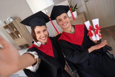 Male graduate in wheelchair with diplomas and his classmate taking selfie at home