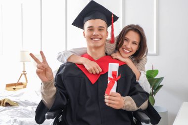 Male graduate in wheelchair with diploma showing victory gesture and his friend at home