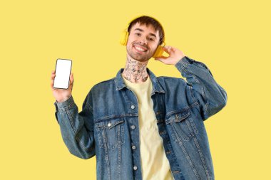 Young tattooed man in headphones with mobile phone listening to music on yellow background