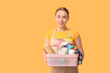 Young woman holding basket with cleaning supplies on orange background