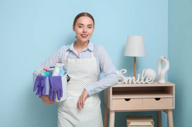 Young woman with cleaning supplies leaning on wooden table near blue wall