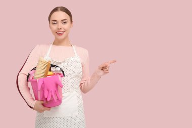 Pretty young woman holding bucket with cleaning supplies and pointing at something on pink background