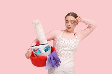 Tired young woman holding basin with cleaning supplies on pink background