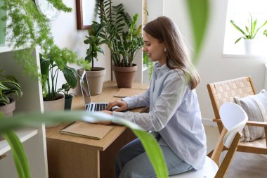 Young woman working with laptop and plants at table in office