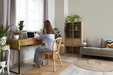 Young woman working with laptop and plants at table near window in office