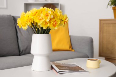 Oturma odasında bir fincan kahve ve dergilerle masasında güzel narsis çiçekleri olan vazo.