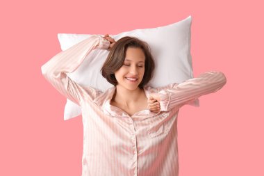 Beautiful young happy sleepy woman in pajamas with pillow on pink background