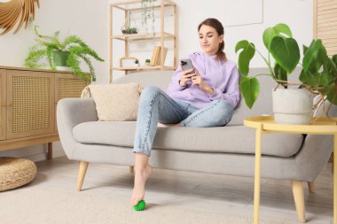 Young woman with mobile phone massaging her foot on sofa at home