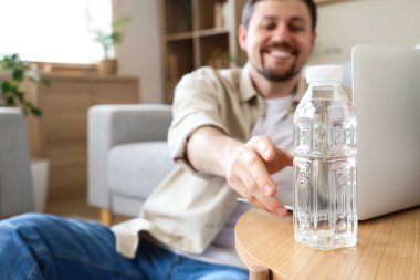 Young man taking bottle of water from table at home, closeup