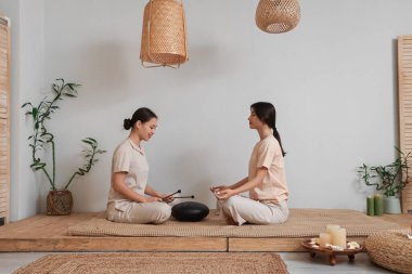 Thai therapist playing glucophone with meditating young woman in spa salon