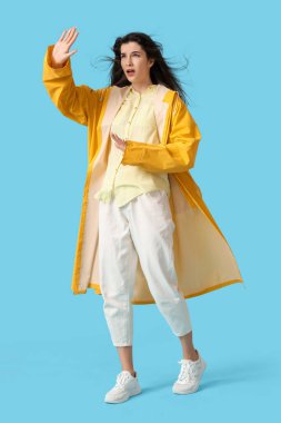 Young woman in raincoat with strong wind on blue background