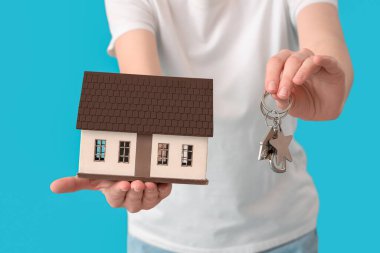 Woman with house model and keys on blue background, closeup. Mortgage concept