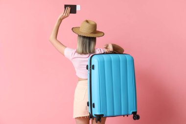 Beautiful young woman with blue suitcase and passport on pink background, back view. Travel concept