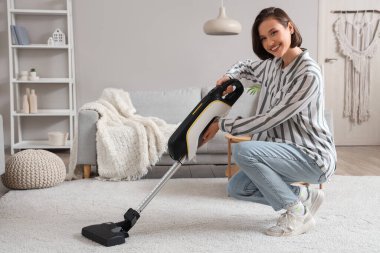 Happy young woman cleaning carpet with handheld vacuum cleaner in living room