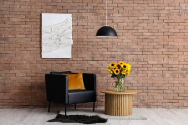 Masada ayçiçekli vazo ve tuğla duvarın yanında siyah koltuk.