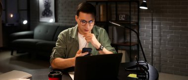 Cep telefonu ve dizüstü bilgisayarı olan yakışıklı bir adam akşam geç saatlerde ofiste.