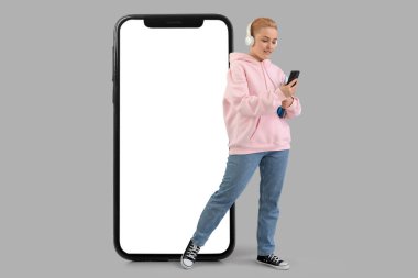 Büyük cep telefonu ve gri arka planda kulaklıkları olan genç bir kadın.