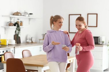 Akıllı telefonlu genç kadınlar mutfakta su içiyorlar.