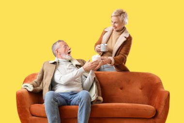 Kakaolu olgun bir çift, sarı arka planda, kanepede oturuyor.