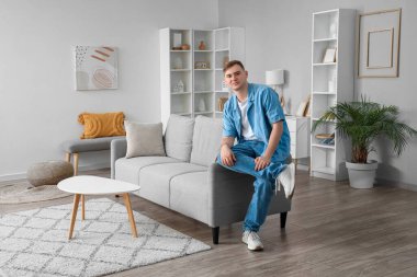 Oturma odasındaki gri koltukta oturan genç adam.