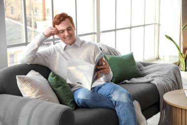 Genç adam oturma odasının penceresinin yanındaki yumuşak koltukta dergi okuyor.