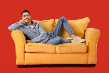 Yakışıklı genç adam, kırmızı arka planda kanepede dinleniyor.