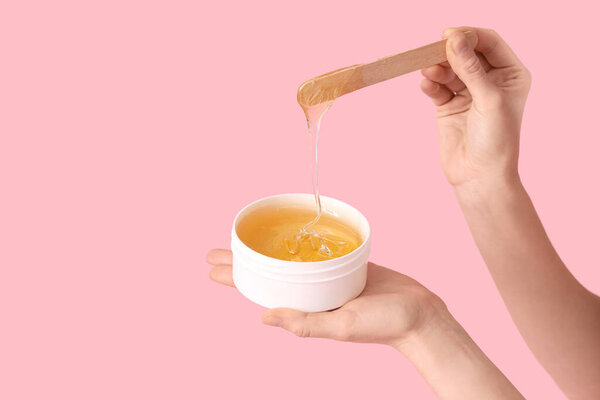 Female hands holding container with sugaring paste and spatula on pink background