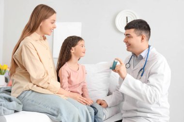 Erkek çocuk doktoru küçük bir kıza ve annesine yatak odasında astım spreyini gösteriyor.